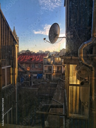 A downward view from an upper window over a dense, cluttered Batumi residential courtyard and rooftop, framed by a window, satellite dish, and utility pipes.