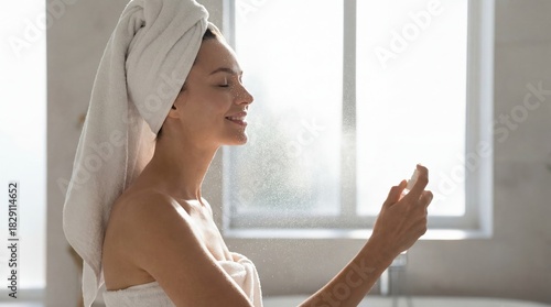 A radiant young woman with a white towel on her head and body gently sprays a refreshing mist onto her smiling face in a brightly lit bathroom.