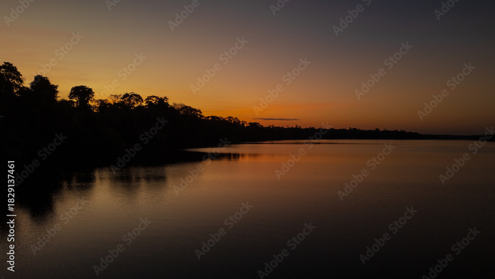 Fototapeta premium Sunset at Lake Valencia with warm orange light reflecting on calm Amazon waters, surrounded by the lush rainforest of Madre de Dios