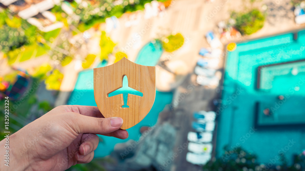 Obraz premium Hand holding wooden shield with airplane icon over blurred resort background, symbolizing travel insurance, vacation protection, flight safety, travel planning, secure trip, and holiday security.