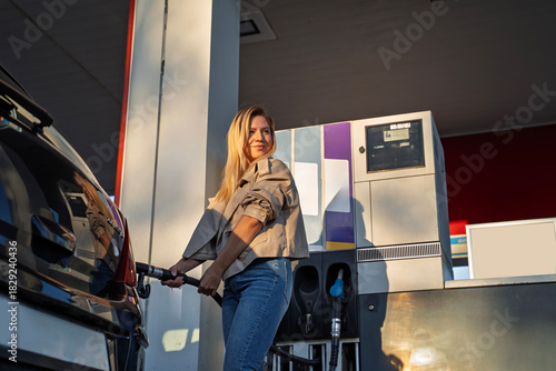 Attractive blonde woman in leather jacket and jeans using fuel nozzle on gas station, refueling her car, beautiful sunset 