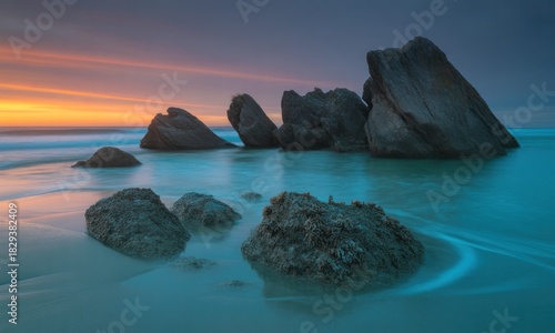 Coastal rocks at sunset.  Turquoise water laps gently around dark rocks on a sandy beach.  Vibrant colors of the sky blend from orange to pink and purple