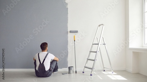 Person sitting on floor admiring freshly painted wall with roller and ladder nearby