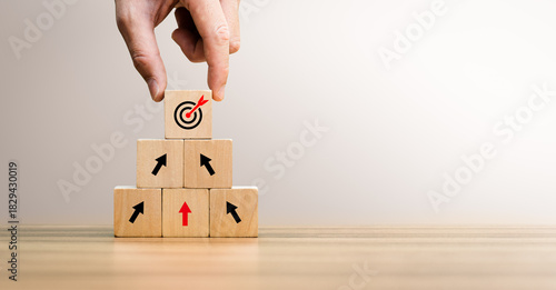 Business growth concept with hand stacking wooden blocks upward, showing arrows and target icon. Represents strategy, goal achievement, development, planning, leadership, and long-term success.