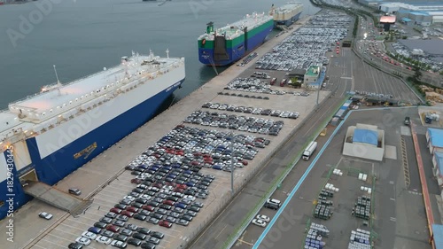 Aerial view of a busy port terminal with two large car carrier ships docked, surrounded by vast parking lots filled with new cars ready for global export, showcasing automotive logistics and trade.