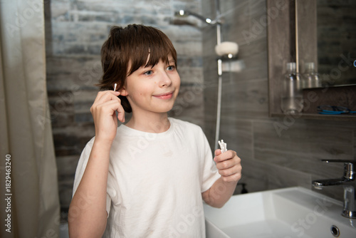 A young boy cleaning his ear with a cotton swab at a bathroom sink as part of a daily grooming routine.