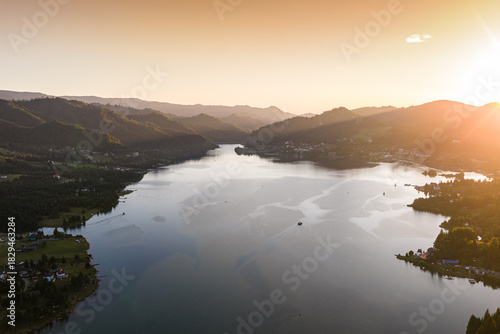 Aerial view of Colibița Lake in the Carpathian Mountains of Romania during a warm summer sunset. Golden light over the water and surrounding hills. Travel to Romania, Transylvania.
