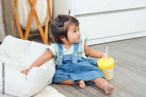 Sweet asian toddler girl sitting on floor leaning pillow holding training cup during relaxing home time symbolizing independence comfort early childhood development and joyful calm lifestyle moment