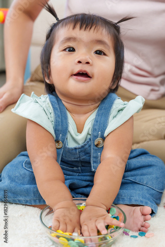 Adorable asian toddler girl sitting on floor playing colorful buttons in glass bowl during home learning activity developing fine motor skill curiosity creativity joyful early childhood growth moment