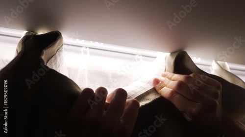 Female hands carefully hanging a thick, heavy curtain on a modern ceiling track against a backlit window, meticulously attaching each hook to the rail for a perfect interior design finish
