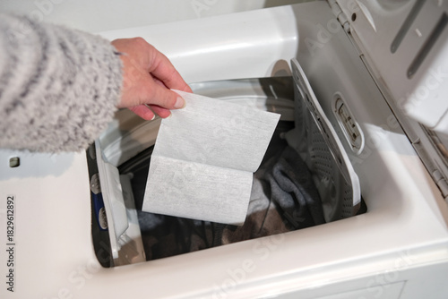 Canvastavla A person holds a color catcher sheet above a top loading washer filled with laundry, preparing to prevent dye transfer during the wash cycle