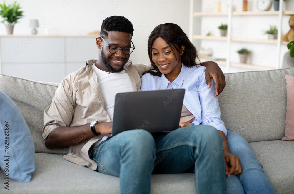 Fototapeta premium Joyful African American couple sits on a sofa at home, enjoying a movie together on a laptop. They cuddle closely, sharing smiles while browsing the internet during their weekend.