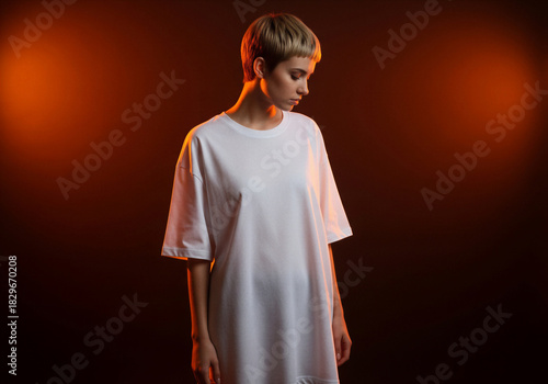 Stylish young woman with short hair wearing white oversized t-shirt in studio, looking down with dramatic moody orange lighting against dark background.