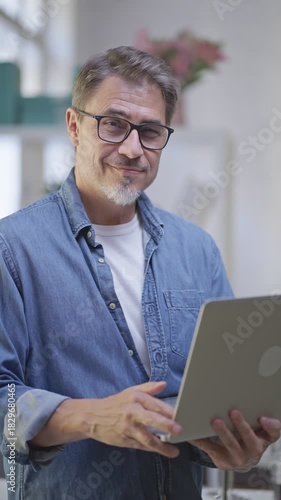 Portrait of middle aged man standing k in bright room working with laptop computer in home office. Mature age, middle age, mid adult casual man in 50s, confident happy smiling.
