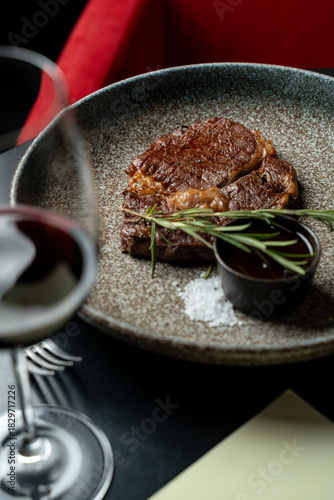 A succulent piece of steak rests on a textured plate beside a small dish of sauce, complemented by a glass of red wine. Fresh rosemary adds a fragrant touch in a cozy restaurant ambiance