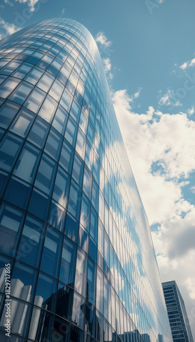 A modern blue glass skyscraper towers into a bright blue sky, reflecting the sky and clouds on its curved facade.
