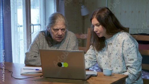 A focused daughter and her concerned mother check serious news on a laptop screen while sitting at a table. They stare intently at the display in a rustic home setting.