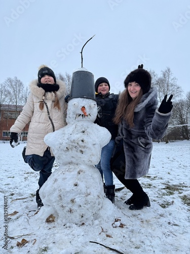 Smiling children and a woman posing with a snowman on a snowy winter day outdoors
