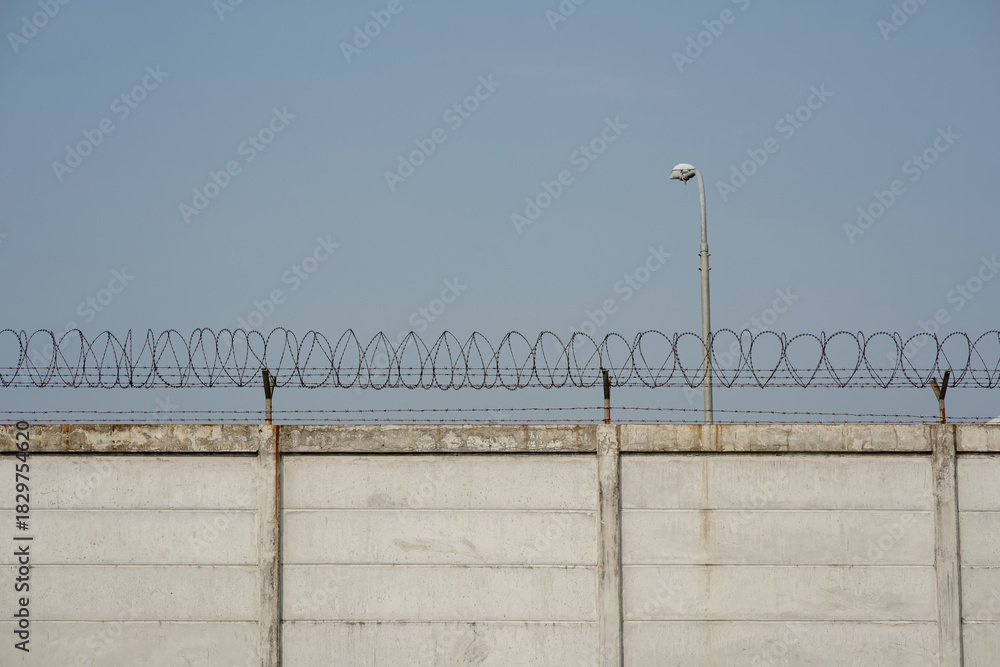 Fototapeta premium Concrete security wall with razor wire fence and street light pole against blue sky background.