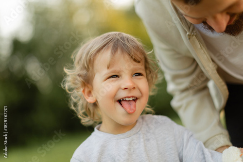 Happy toddler sticking out tongue while playing outdoors, with parent leaning in beside him. Warm natural light, candid family moment full of joy and connection.