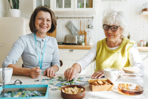 Two cheerful ladies playing puzzle together at kitchen table, mature female therapist working with senior patient with dementia using method of memory stimulation through board games