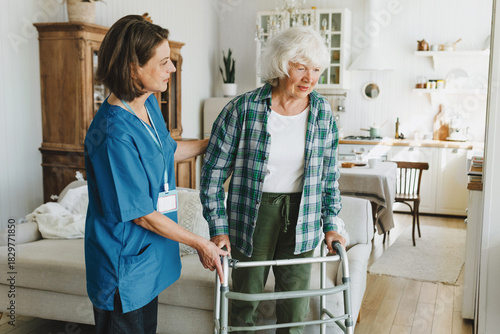 Side view of female caregiver in blue uniform with badge assisting elderly woman with arthritis walking with mobility walker in living-room within physical rehabilitation treatment program