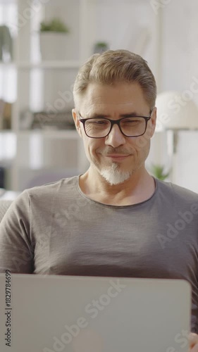 Older man working online with laptop computer at home sitting on couch in living room. Home office, browsing internet. Portrait of happy, mature age, middle age, mid adult man in 50s, smiling.