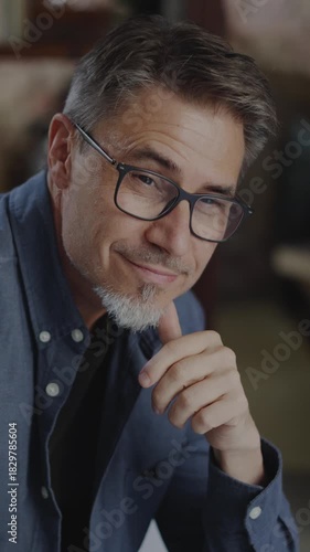 Confident older white man at home, sitting on couch in dark living room. Portrait of happy, mature age, middle age, mid adult caucasian man in 50s, smiling, looking at camera.