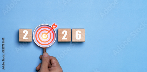 Hand holding magnifying glass highlighting a target icon between the numbers 2026 represents a happy new year with focus goal setting motivation precision planning and direction for future success.