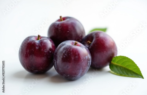 Four fresh ripe purple plums arranged on pure white background. One vibrant green leaf rests beside delicious fruit. Sweet berries offer healthy nutrition. Perfect for desserts, snacks, balanced diet.