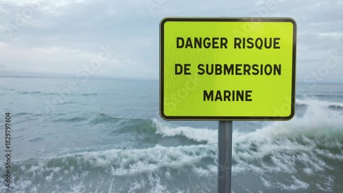 Slow motion turquoise sea storm waves crashing against a seawall, spraying foam around a danger marine submersion sign.