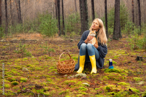 Woman in yellow boots sitting on moss in the forest and holding a ginger cat on her knees. Basket and thermos beside her. Cozy outdoor moment with pet and nature.