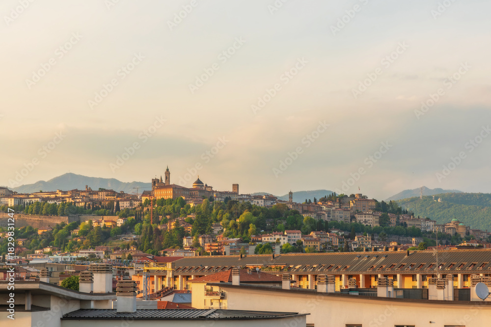 Fototapeta premium Panoramic view of Bergamo old town at sunset with historical buildings and lush green hills in the background, Lombardy, Italy. Citta alta medieval skyline with its towers, domes