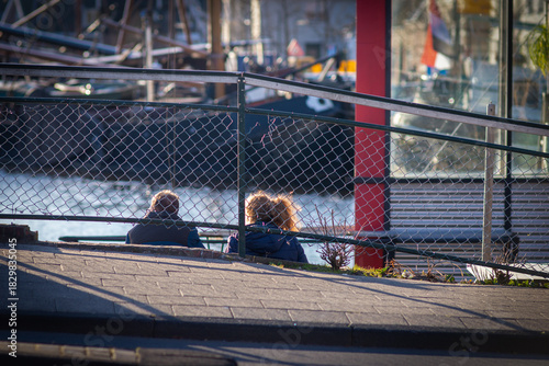 Wallpaper Mural Quiet afternoon by the Leiden canal where two figures sit behind a chain-link fence, sunlight glinting on water, boats and glass as early spring warmth softens the city in calm March light! Torontodigital.ca