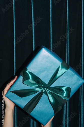Woman's hands holding blue gift box behind green velvet curtain
