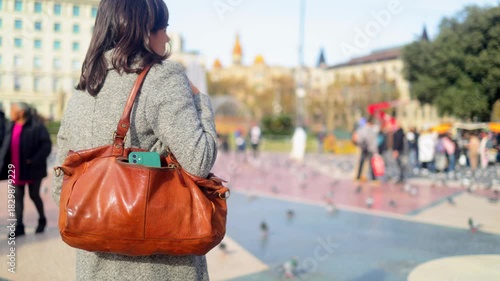Woman walking through a busy barcelona city square, her handbag open with a mobile phone visible, symbolizing the risk of theft and the importance of traveler caution in an urban environment