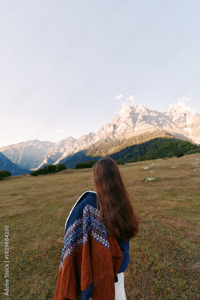 Naklejka premium Woman back view standing in meadow, looking at snow-capped mountains and wide landscape. Nature travel scene with long hair, coat draped over shoulder and hiking mood.