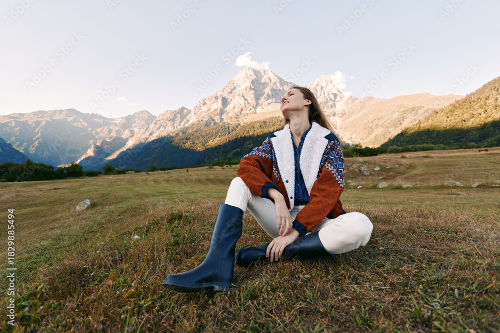 Fototapeta premium Woman mountains meadow boots sweater travel hiking, relaxed portrait of a young woman sitting in a grassy alpine meadow wearing knit sweater and boots, enjoying scenic mountain landscape.