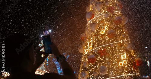 A person captures the magic of a festive Christmas tree adorned with sparkling lights and musical notes, surrounded by falling snow at night. Celebrating winter holidays and