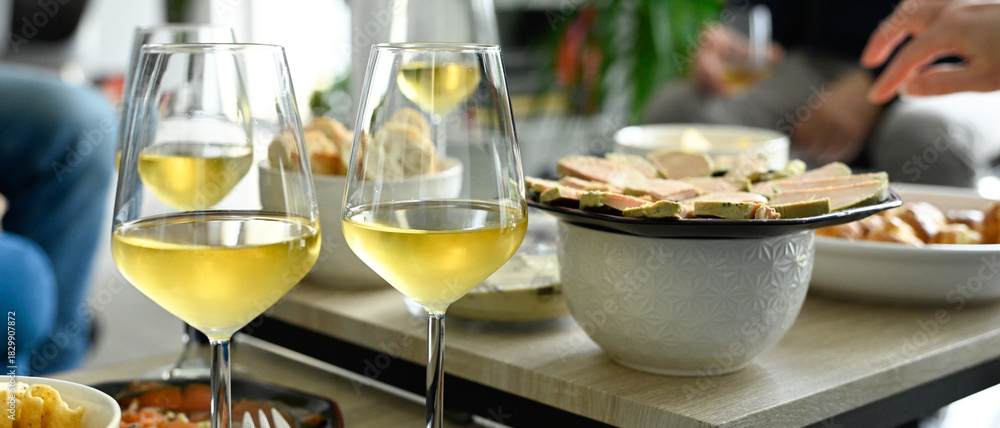 Naklejka premium Scène d'apéritif composé de plusieurs verres de vin blanc et d'assiettes de foie gras et saumon fumé, sur des tables basses dans un salon de jour, format bannière