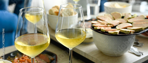 Scène d'apéritif composé de plusieurs verres de vin blanc et d'assiettes de foie gras et saumon fumé, sur des tables basses dans un salon de jour, format bannière