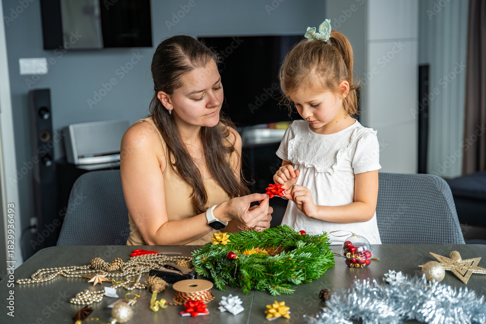 Fototapeta premium Mother and her little daughter decorating a Christmas wreath together at home. Strengthening family bonds through shared festive traditions and creative seasonal activities.