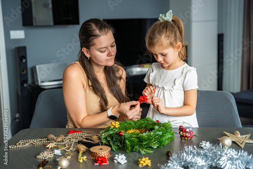 Mother and her little daughter decorating a Christmas wreath together at home. Strengthening family bonds through shared festive traditions and creative seasonal activities.