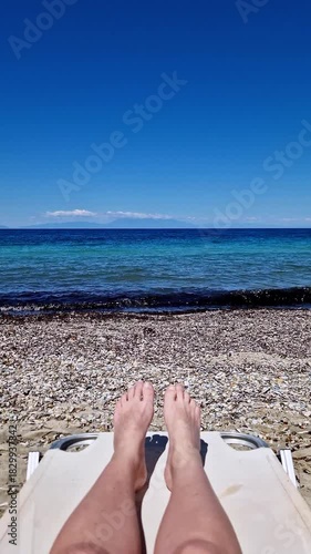 Point-of-view shot of bare legs resting on a white sunbed overlooking a pebble beach. The scene features calm turquoise ocean waves under a clear blue sky.