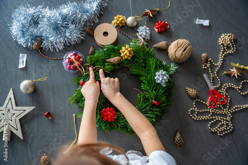 Little girl decorating a Christmas wreath while sitting at the table, top view. Capturing a hands-on festive activity from an overhead perspective at home.