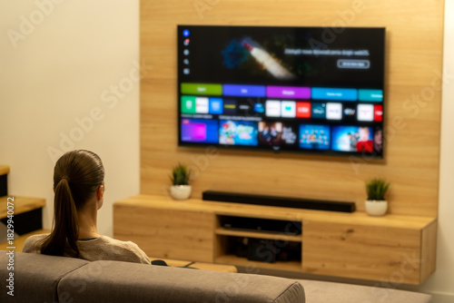 Woman watching smart TV while sitting on sofa in living room. Modern, minimalist interior. Streaming, entertainment, hobbies, free time. LED technology, soundbar