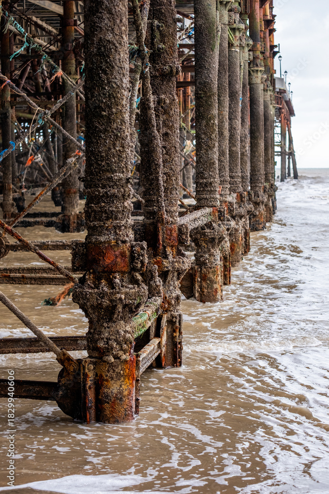 Fototapeta premium Weathered wooden pier supports covered in barnacles stand in shallow water, with gentle waves lapping at the base, creating a serene coastal atmosphere and showcasing natural textures
