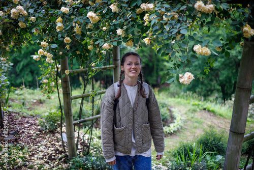 A smiling woman with braids stands beneath a lush arch of pale yellow roses and green leaves, framed by bamboo posts in a sunlit garden.