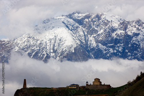 Snow-capped mountains loom above a misty valley, with a stone church and tower perched on a green hillside under overcast skies.