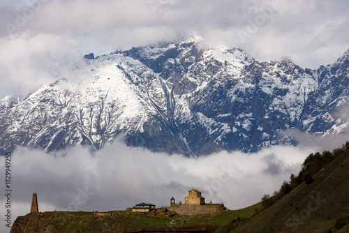 Snow-capped mountains loom above a valley shrouded in clouds, with a stone church and tower perched on a green hillside under soft, diffused light.